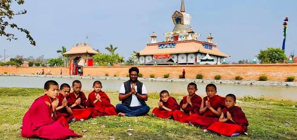 Dr. D.P. Shukla seated on a lawn with young monks in red robes, a temple behind them