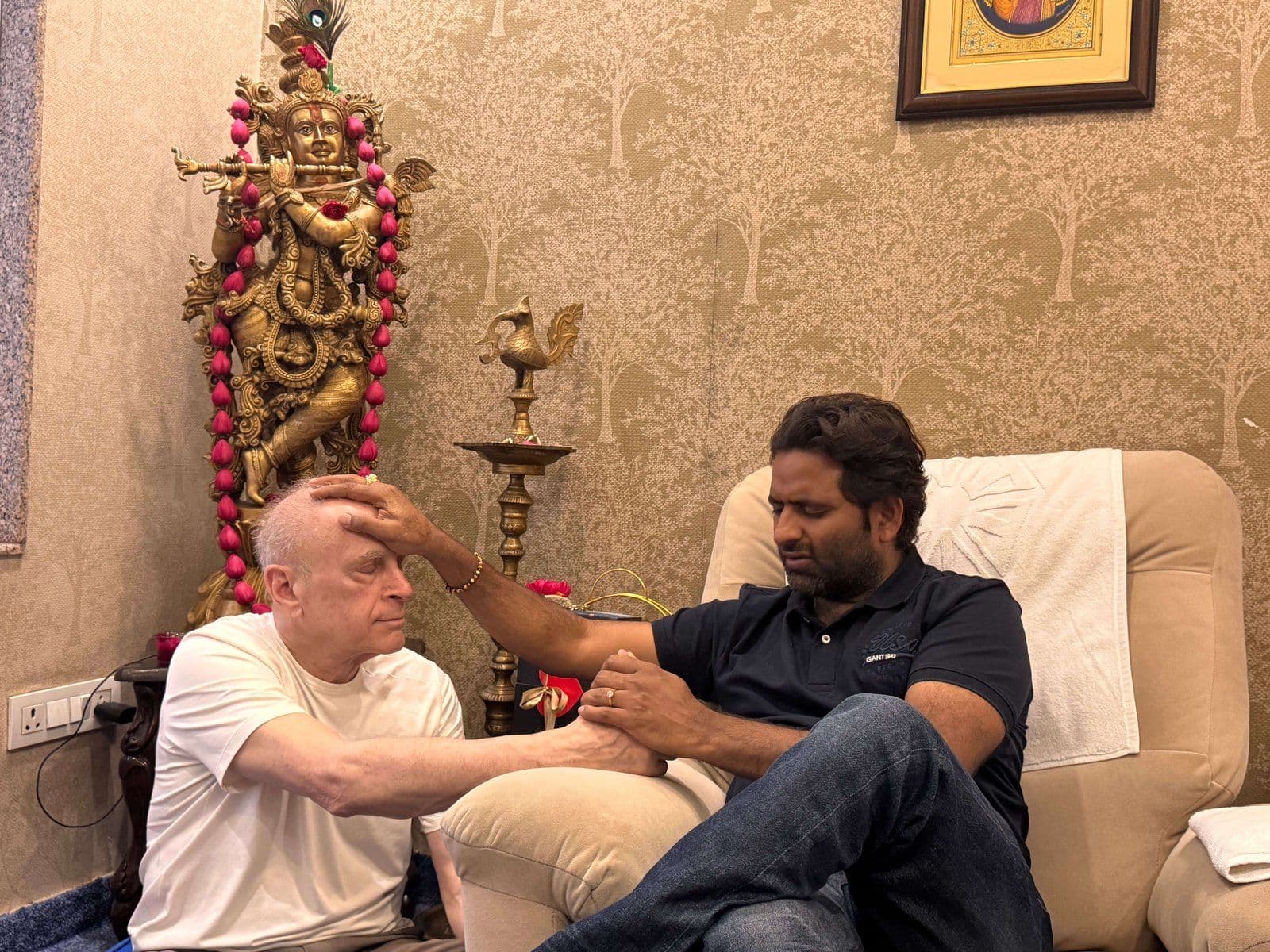 Dr. D.P. Shukla placing a hand on a visitor’s head beside a brass Krishna statue
