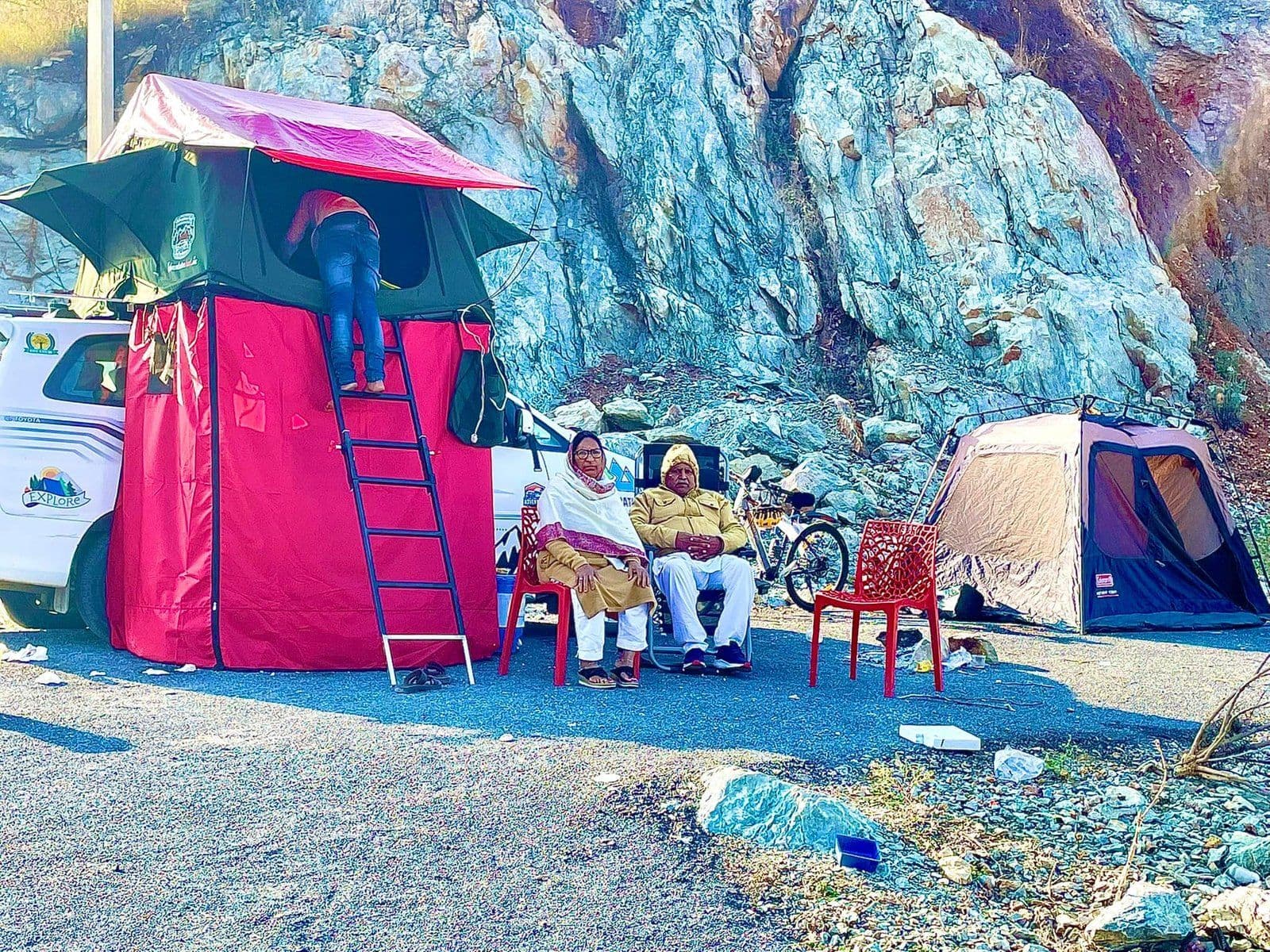 Two people seated beside a rooftop-tent camper against a rock face